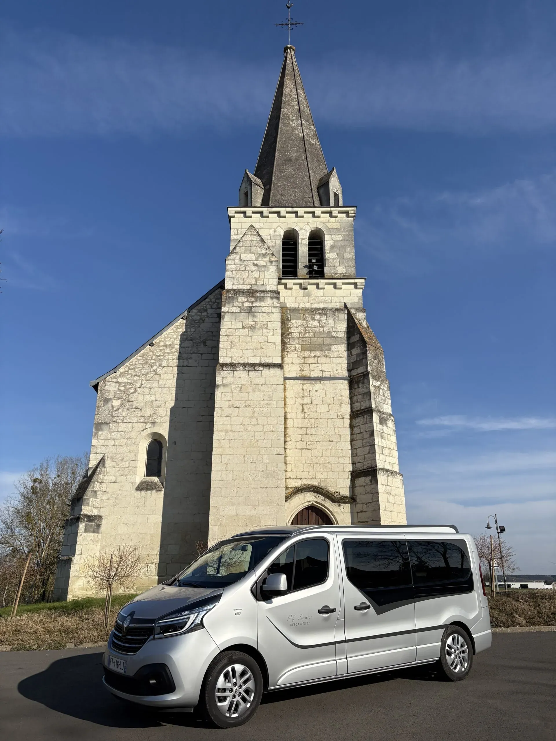Véhicule funéraire gris stationné devant une église en pierre au ciel dégagé.